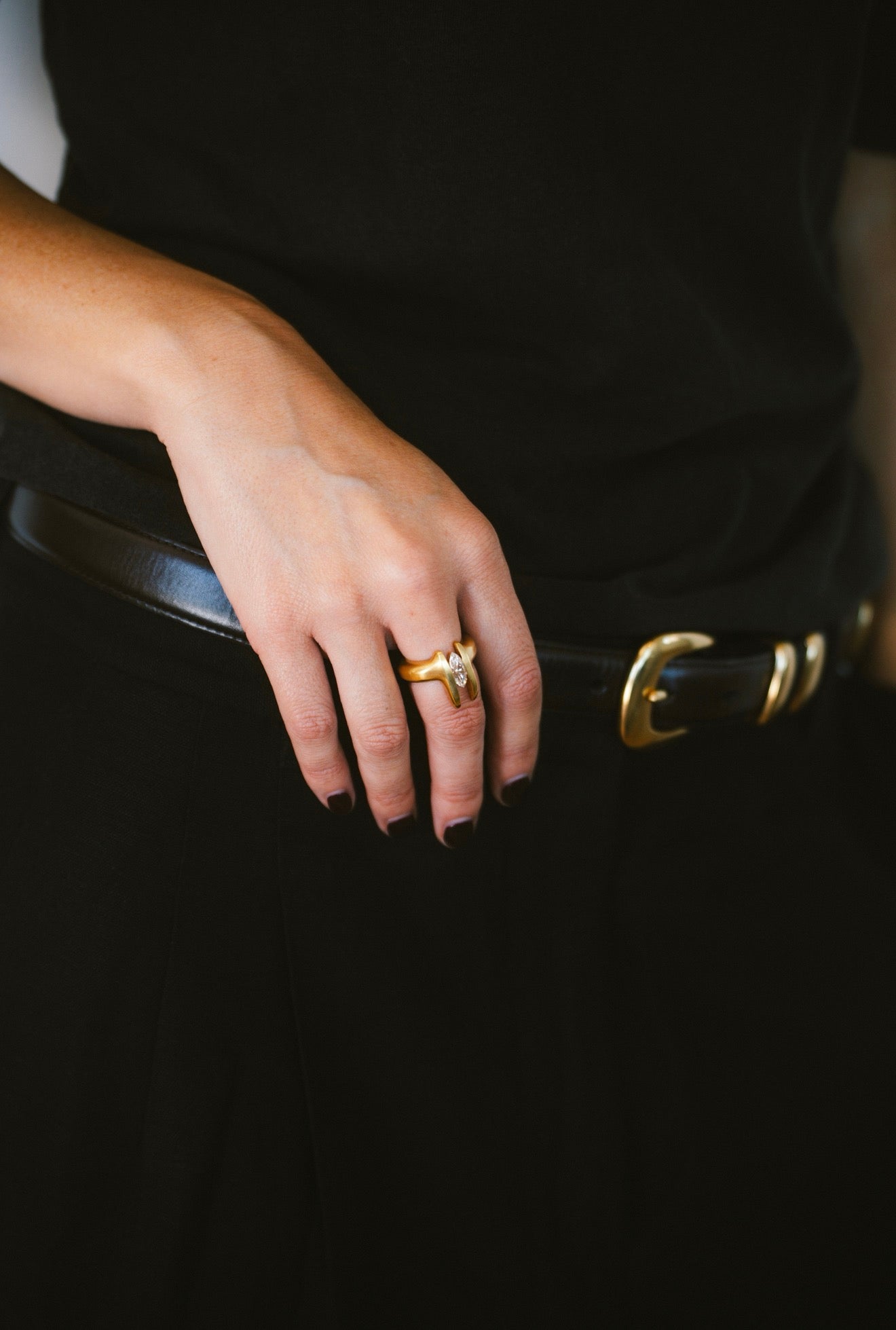 A close-up of a person's hand resting on a black belt with gold-tone hardware, wearing a gold band ring with a small clear center stone.